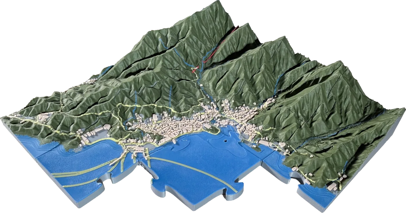 A TopoSaic terrain puzzle of Miyajima, Japan, printed in colour and assembled: a town along a narrow shore below steep forested mountains, with printed roads, streams and individual buildings, and a few pieces lifted out so the interlocking cuts show through the water